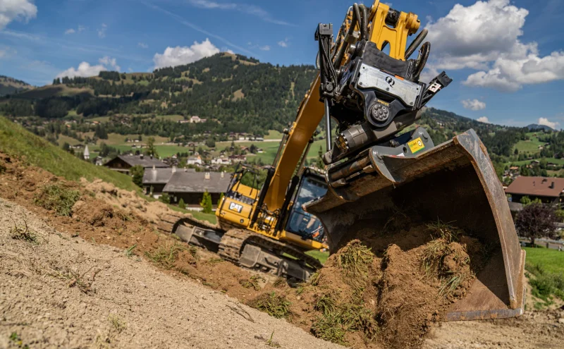 Bagger mit Profillöffel und OilQuick Tiltrotator-Sandwich im Einsatz an einem Hang, der Erdreich und Gras bewegt.