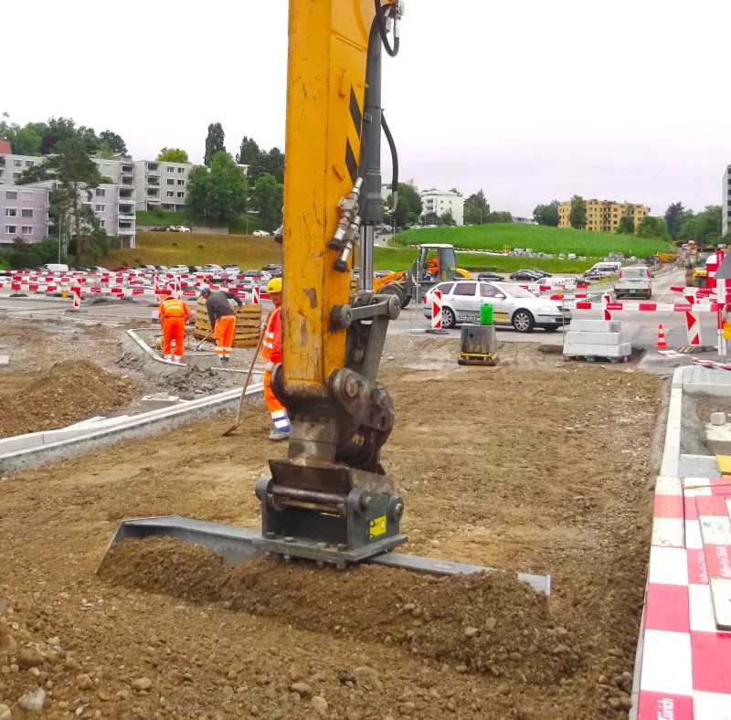 Ein Bagger arbeitet mit einem Planierschild auf einer Straßenbaustelle, während Bauarbeiter im Hintergrund Bordsteine setzen.